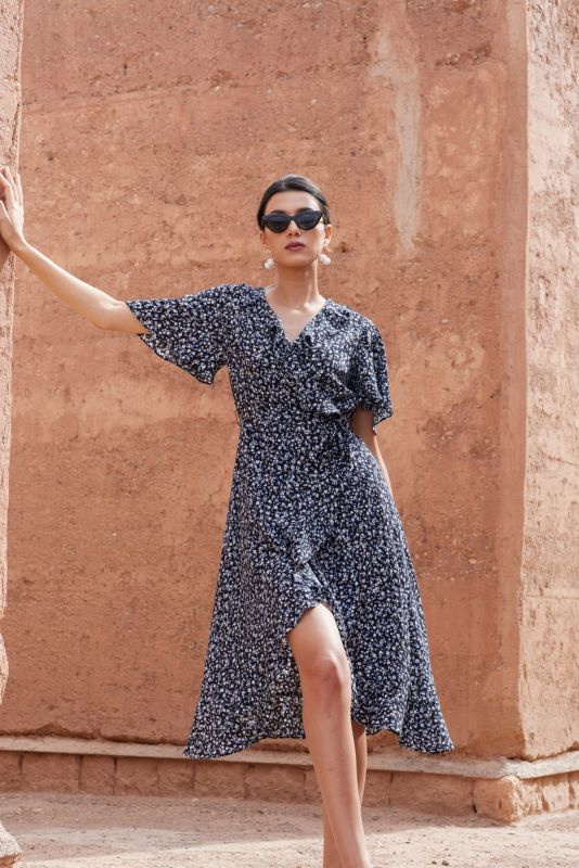 Femme portant une robe noire à petites fleurs blanches et bleues, posant devant un mur en terre cuite.