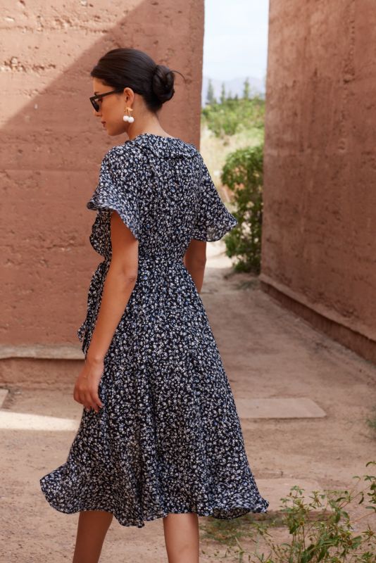 Femme portant une robe noire à motifs floraux blancs, posant contre un mur en terre cuite.