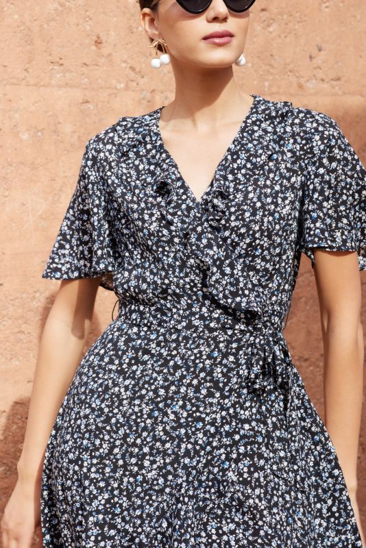 Femme portant une robe noire à petites fleurs blanches et bleues, posant devant un mur en terre cuite.