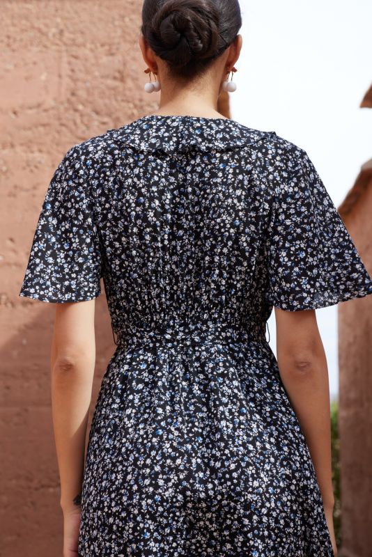 Femme portant une robe noire à petites fleurs blanches et bleues, posant devant un mur en terre cuite.