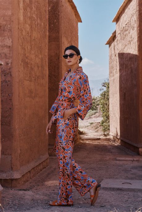 Femme portant un ensemble à motifs floraux rouges, bleus et marron, posant de profil entre deux murs en terre cuite.