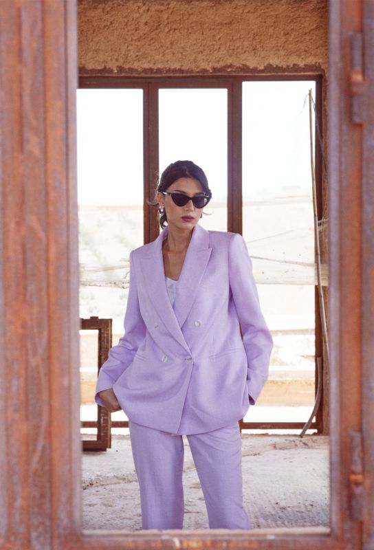 Femme en veste Teresa lilas, posant avec des lunettes de soleil et des boucles d'oreilles, mur en arrière-plan.