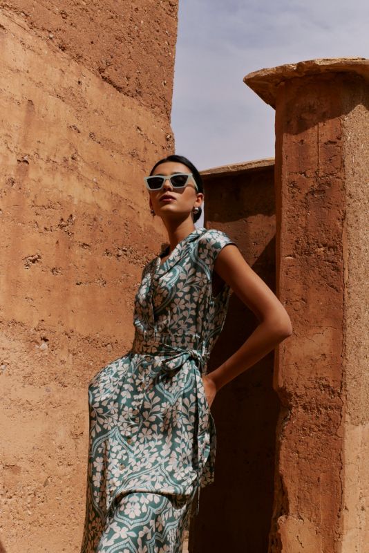 Femme portant la robe Aude à motifs floraux verts, posant contre un mur en terre cuite avec des lunettes de soleil.