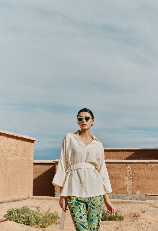 Femme posant avec la blouse Amanda beige et un pantalon vert à motifs, devant un bâtiment en terre cuite.