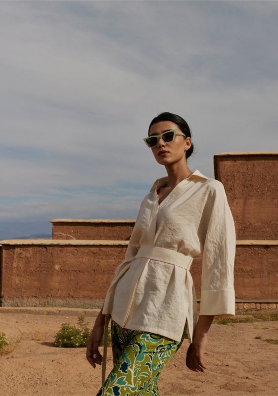Vue latérale de la blouse Amanda beige avec une ceinture, portée par une femme avec des lunettes de soleil, en extérieur.