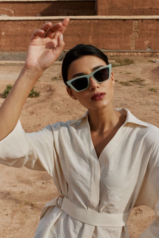 Femme posant avec la blouse Amanda beige et un pantalon vert à motifs, devant un bâtiment en terre cuite.