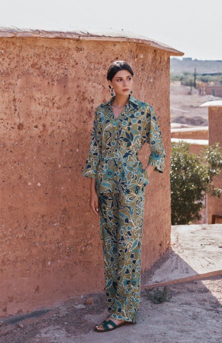 Femme portant le pantalon Romane à motifs floraux verts, avec une chemise assortie, posant devant un mur en terre cuite.