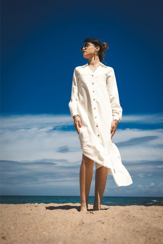 Femme debout sur la plage, portant la robe Marta en lin blanc, avec un ciel bleu en arrière-plan.