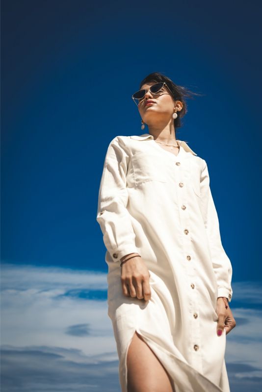 Femme posant de profil sur la plage, portant la robe Marta en lin blanc, sous un ciel bleu clair.