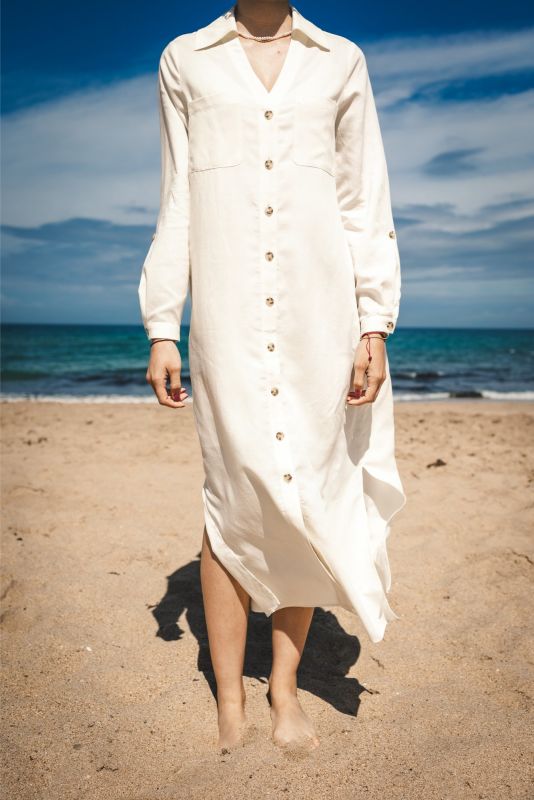 Femme posant sur la plage, portant la robe Marta en lin blanc, les pieds nus dans le sable.
