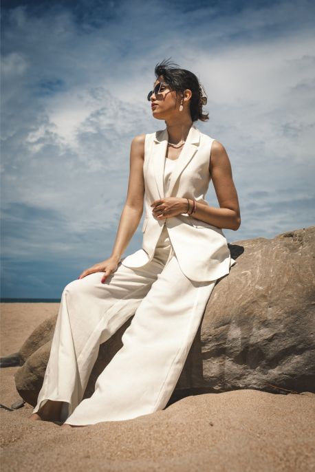 Femme assise sur un rocher à la plage, portant le pantalon Blanca en lin blanc et un gilet assorti.